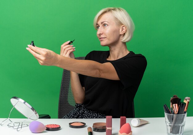 Feliz joven hermosa se sienta a la mesa con herramientas de maquillaje sosteniendo rímel en el lado aislado en la pared verde