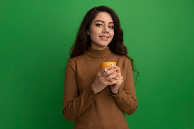 Feliz joven hermosa sosteniendo una taza de té aislado en la pared verde con espacio de copia