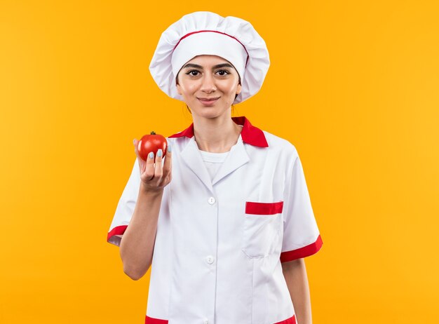 Feliz joven hermosa en uniforme de chef con tomate aislado en la pared naranja