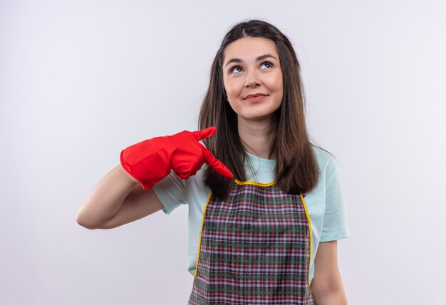 Feliz joven hermosa vestida con delantal y guantes de goma apuntando con el dedo índice a sí misma