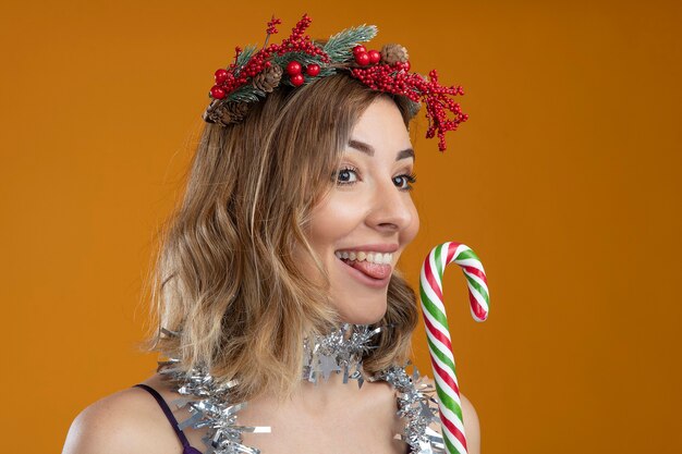 Feliz joven hermosa vestida de púrpura con corona con guirnalda en el cuello sosteniendo dulces navideños aislado sobre fondo marrón