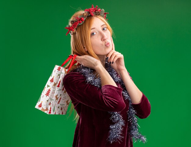 Feliz joven hermosa vestida de rojo con corona y guirnalda en el cuello sosteniendo una bolsa de regalo en el hombro aislado sobre fondo verde