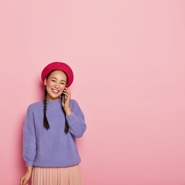 Feliz joven japonesa se relaja durante la conversación telefónica, habla de algo agradable, viste ropa brillante