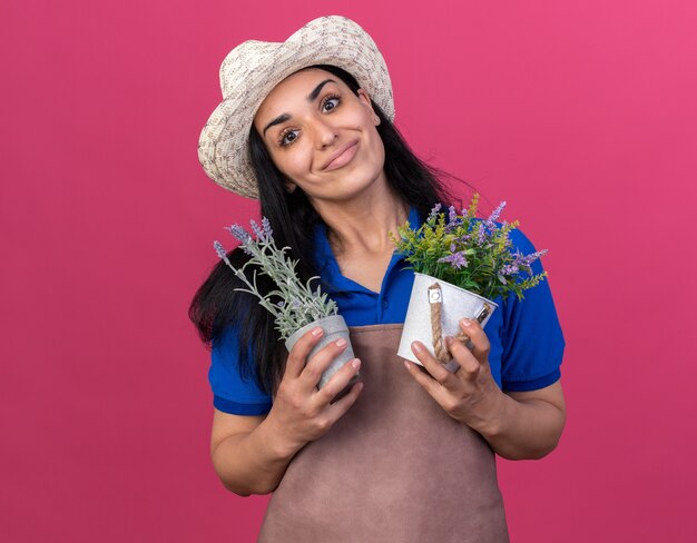 Feliz joven jardinero caucásico vestida con uniforme y sombrero sosteniendo macetas aisladas en la pared rosa con espacio de copia