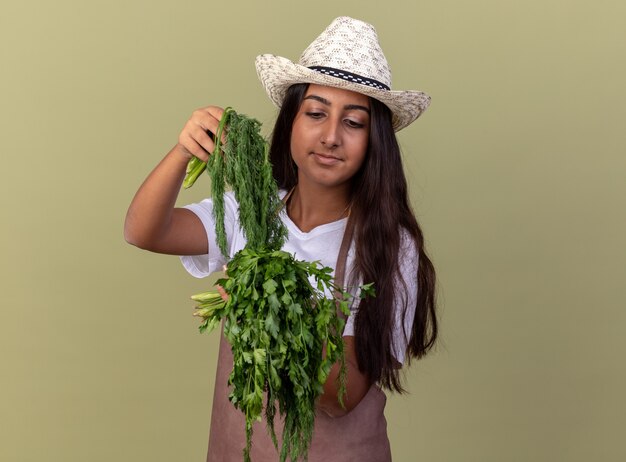 Feliz joven jardinero chica en delantal y sombrero de verano con hierbas frescas sonriendo confiado en la cara de pie sobre la pared verde