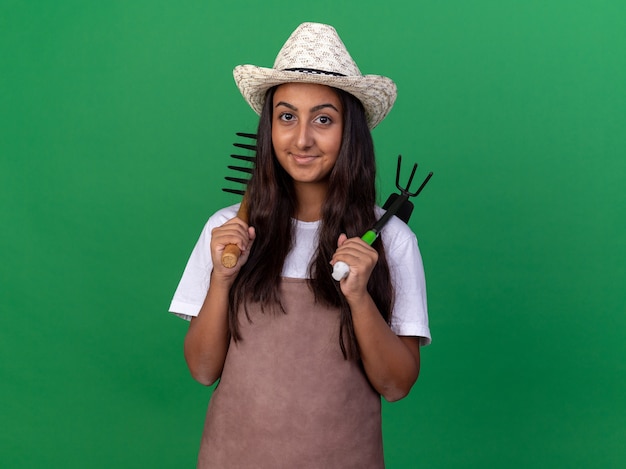 Feliz joven jardinero con delantal y sombrero de verano sosteniendo mini rastrillo y azadón con una sonrisa en la cara de pie sobre la pared verde