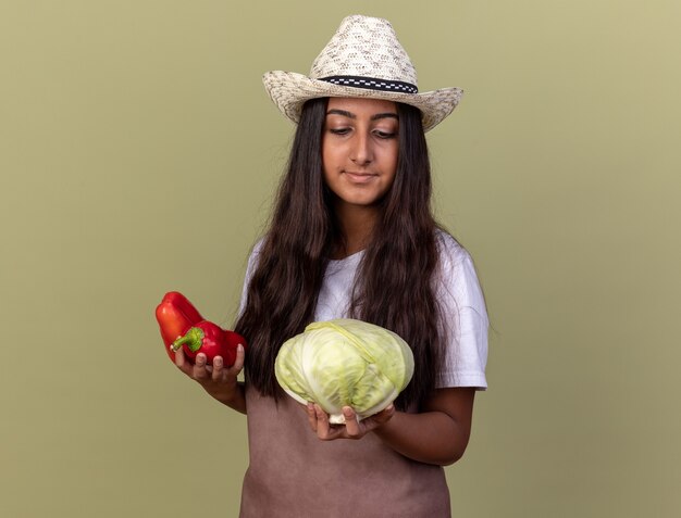 Feliz joven jardinero con delantal y sombrero de verano sosteniendo pimientos rojos frescos y repollo mirando confiado sonriendo de pie sobre la pared verde