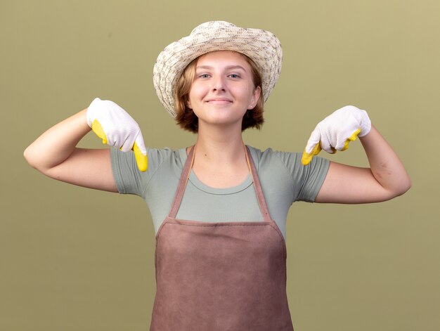 Feliz joven jardinero eslavo vistiendo guantes y gorro de jardinería apuntando hacia abajo en verde oliva