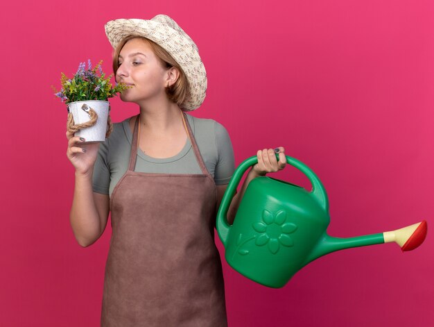 Feliz joven jardinero eslavo vistiendo sombrero de jardinería sosteniendo regadera y oliendo flores en maceta en rosa