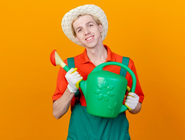 Feliz joven jardinero hombre vestido con mono y sombrero sosteniendo regadera mirando a la cámara sonriendo alegremente de pie sobre fondo naranja