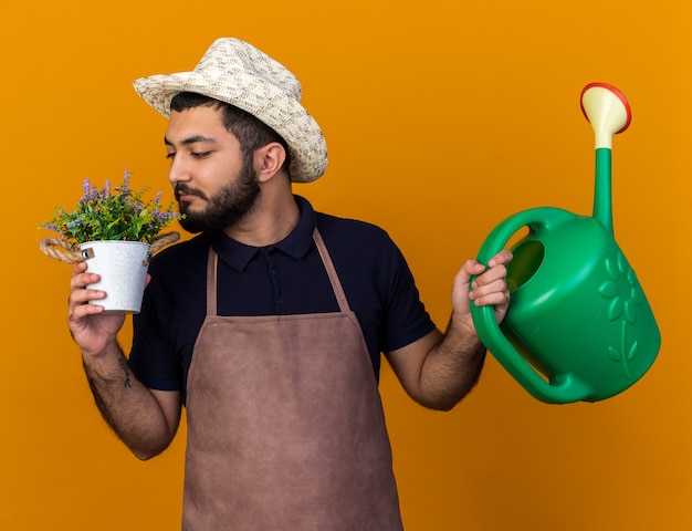 Foto gratuita feliz joven jardinero varón caucásico con sombrero de jardinería sosteniendo regadera y oliendo flores en maceta aislado en la pared naranja con espacio de copia