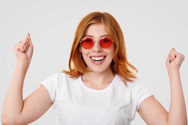 Feliz joven jengibre exitosa hembra mantiene los puños cerrados levantados, tiene una sonrisa de dientes, celebra el día exitoso