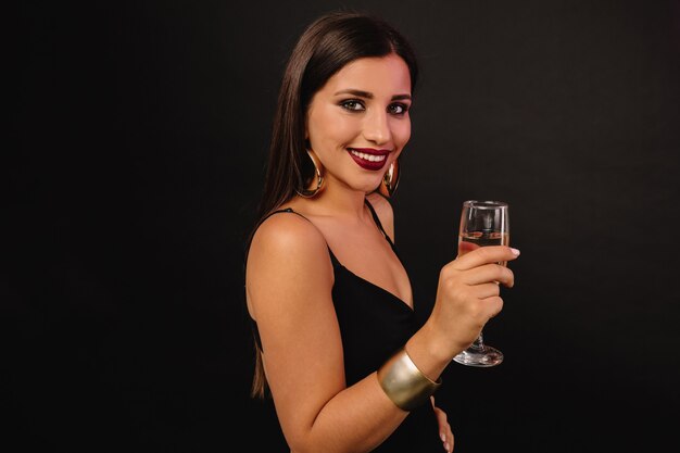 Feliz joven con joyas de oro en vestido negro bebiendo champán