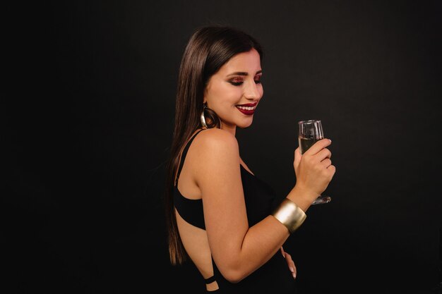 Feliz joven con joyas de oro en vestido negro bebiendo champán