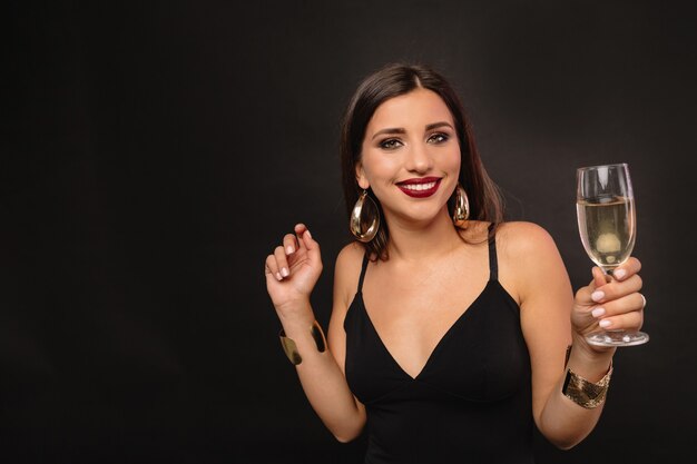 Feliz joven con joyas de oro en vestido negro bebiendo champán