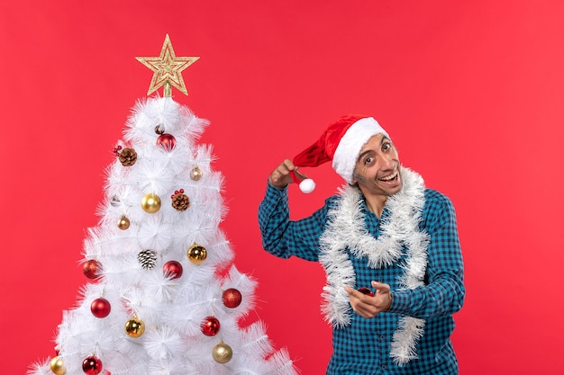 Feliz joven jugando con sombrero de santa claus en una camisa azul a rayas y árbol de Navidad en rojo