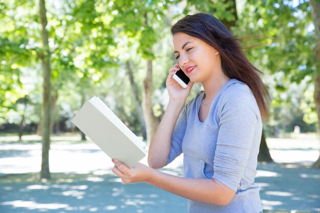 Feliz joven leyendo libro y llamando por teléfono en el parque