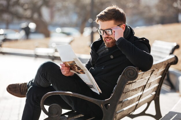 Feliz joven leyendo el periódico en el banco de la ciudad