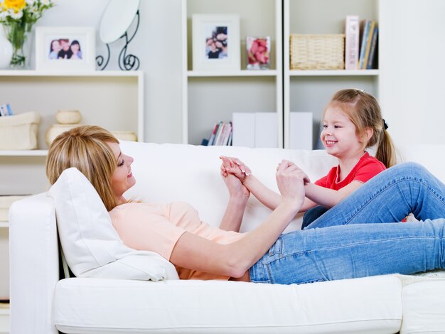 Feliz joven madre relajándose en el sofá y jugando con su pequeña hija - en el interior