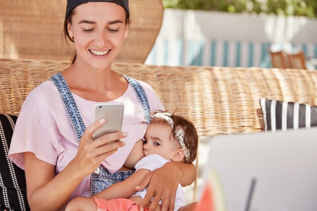 Feliz joven madre sonriente en ropa casual amamanta a su pequeño bebé, feliz de recibir mensajes de texto en el teléfono inteligente, ama a su hijo, hace compras en línea. Concepto de maternidad y maternidad