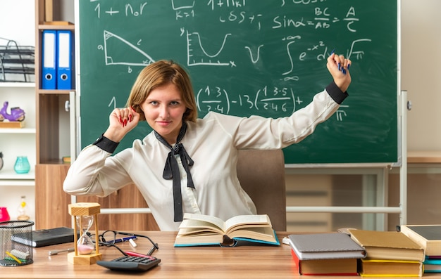 Feliz joven maestra se sienta a la mesa con útiles escolares en el aula