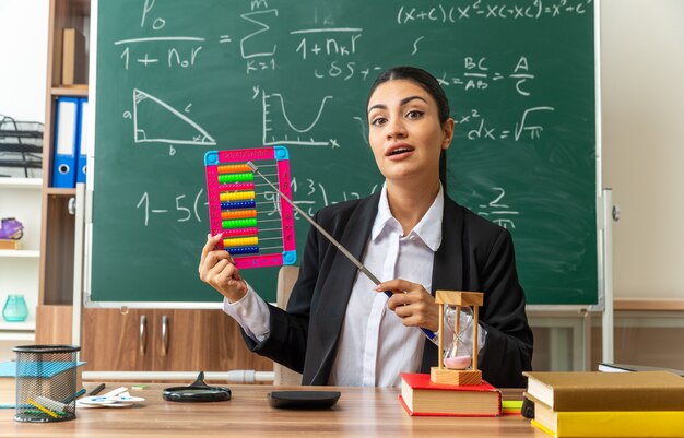 Feliz joven maestra se sienta a la mesa con útiles escolares puntos en el ábaco con puntero en el aula