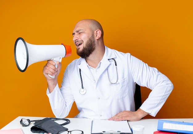 Feliz joven médico calvo vestido con bata médica y estetoscopio sentado en la mesa de trabajo con herramientas médicas habla por altavoz aislado sobre fondo naranja