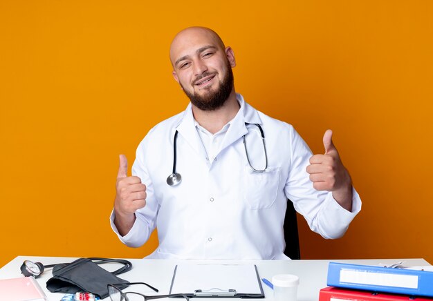 Feliz joven médico varón calvo vistiendo bata médica y estetoscopio sentado en el escritorio con herramientas médicas sus pulgares arriba aislados sobre fondo naranja