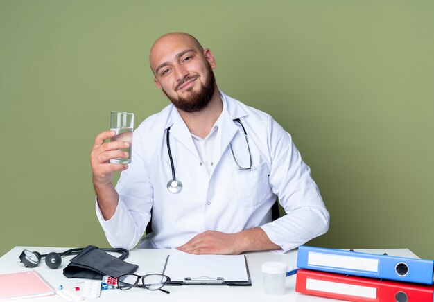 Feliz joven médico varón calvo vistiendo bata médica y estetoscopio sentado en el escritorio de trabajo con herramientas médicas sosteniendo un vaso de agua aislado sobre fondo verde
