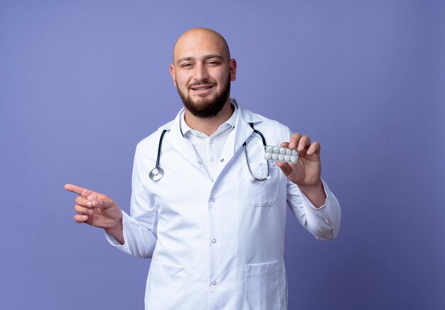 Feliz joven médico varón calvo vistiendo bata médica y estetoscopio sosteniendo píldoras y puntos en el lado aislado sobre fondo azul con espacio de copia