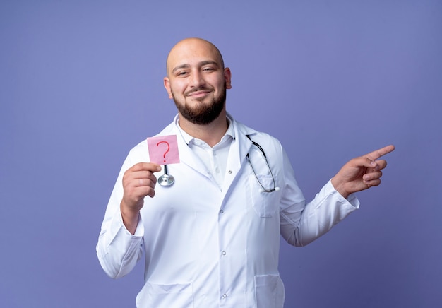 Feliz joven médico varón calvo vistiendo una bata médica y un estetoscopio sosteniendo un signo de interrogación de papel y puntos al costado