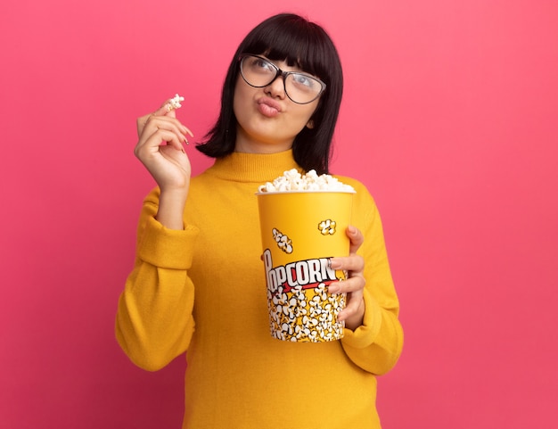 Feliz joven morena caucásica con gafas ópticas sostiene un cubo de palomitas de maíz y mira a un lado