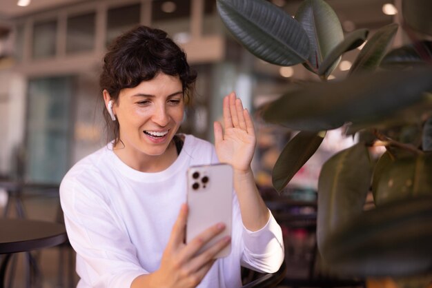 Feliz joven morena caucásica con ropa informal y chats de auriculares con amigos en un teléfono inteligente sentado en un café