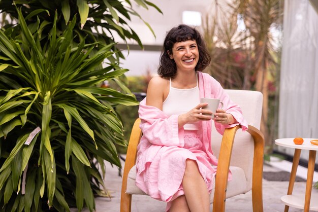 Feliz joven morena caucásica sonriendo a la cámara mientras se sienta en el patio con una taza de café en la mañana Concepto de estilo de vida