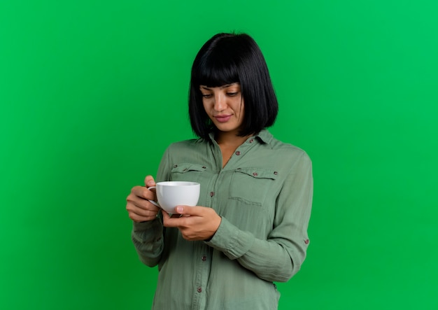 Foto gratuita feliz joven morena caucásica sostiene y mira la taza aislada sobre fondo verde con espacio de copia