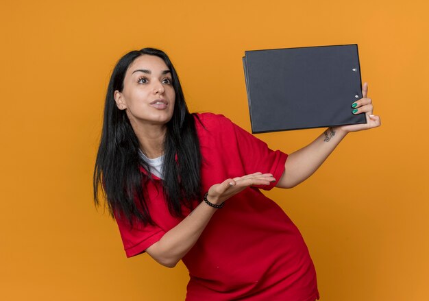 Feliz joven morena caucásica vestida con camisa roja sostiene y apunta al portapapeles con la mano aislada en la pared naranja