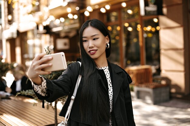 Feliz joven morena en elegante gabardina negra sonríe sinceramente, sostiene el teléfono y toma selfie afuera cerca de la cafetería de la calle