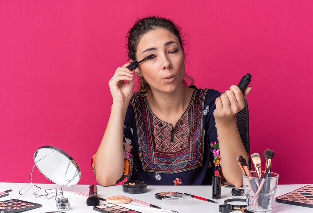 Feliz joven morena sentada a la mesa con herramientas de maquillaje aplicando rímel aislado en la pared rosa con espacio de copia