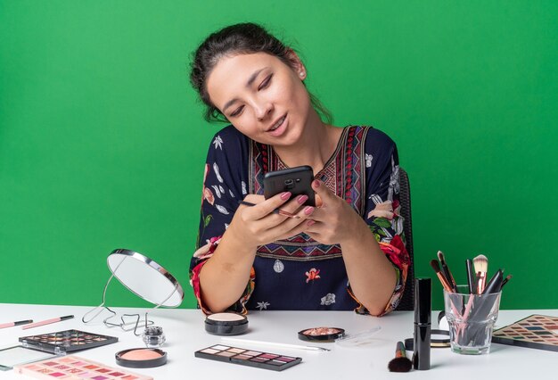 Feliz joven morena sentada a la mesa con herramientas de maquillaje sosteniendo y mirando el teléfono aislado en la pared verde con espacio de copia