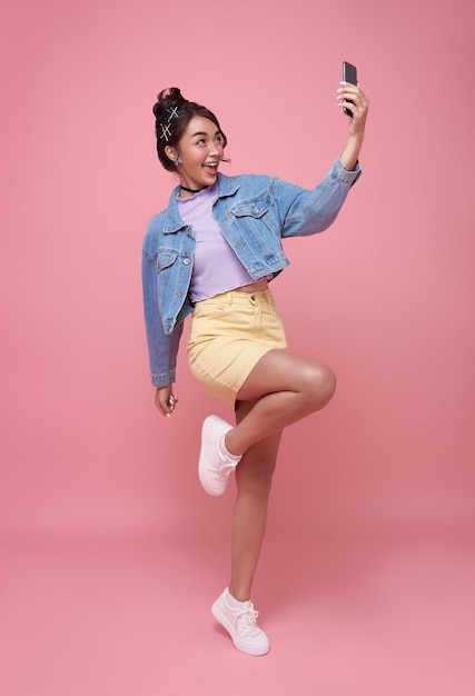 Feliz joven mujer adolescente asiática celebrando con teléfono móvil aislado sobre fondo rosa.