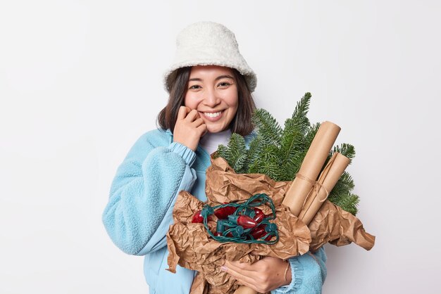 Feliz joven mujer asiática usa sombrero de invierno y chaqueta azul mira tiernamente a la cámara tiene atributos de Año Nuevo para decorar la casa disfruta de sonrisas de vacaciones alegremente aisladas sobre fondo blanco