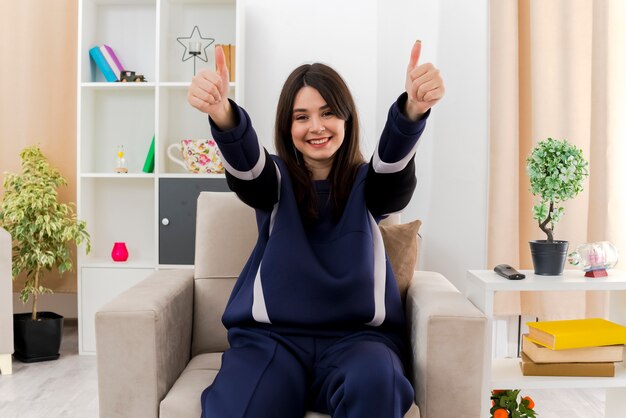 Feliz joven mujer bonita caucásica sentada en un sillón en la sala de estar diseñada mostrando los pulgares hacia arriba y mirando