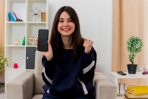 Feliz joven mujer bonita caucásica sentada en un sillón en la sala de estar diseñada que muestra el teléfono móvil mirando y apretando el puño