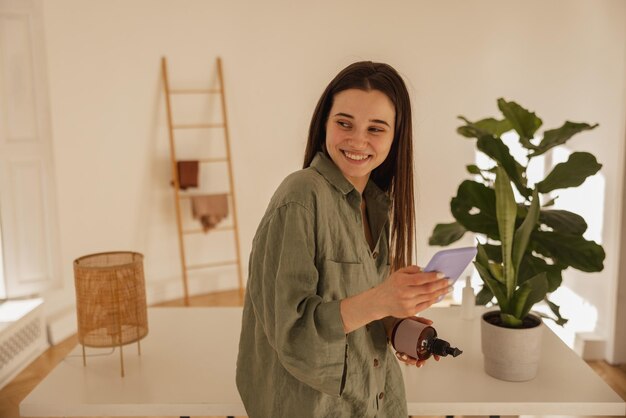 Feliz joven mujer caucásica sosteniendo un teléfono móvil mirando hacia otro lado mientras está de pie en la habitación Chica morena lee un mensaje de amor en un chat personal con una sonrisa en la cara Concepto de disfrutar el momento