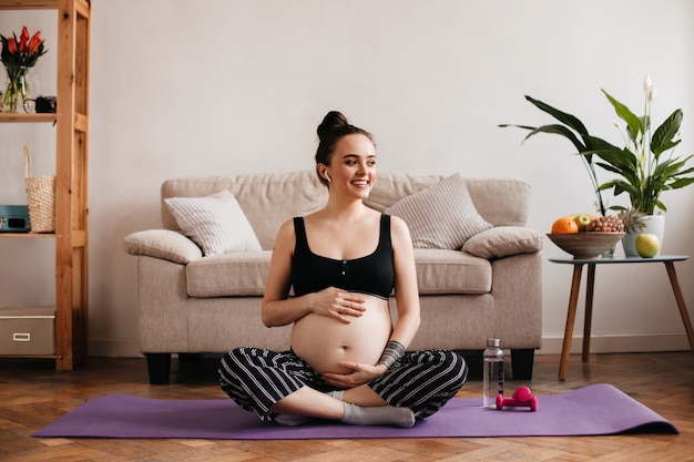 Foto gratuita feliz joven mujer embarazada sonríe y toca suavemente el vientre. chica morena en top negro y pantalones a rayas se sienta en la alfombra de yoga y escucha música en auriculares inalámbricos.