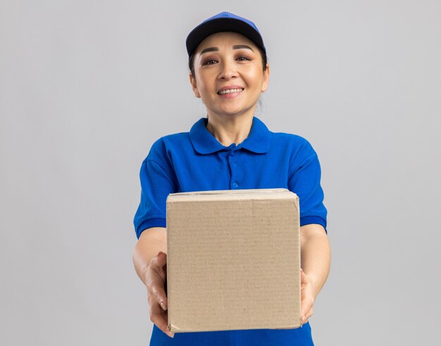 Feliz joven mujer de entrega en uniforme azul y gorra sosteniendo una caja de cartón con una sonrisa en la cara de pie sobre la pared blanca