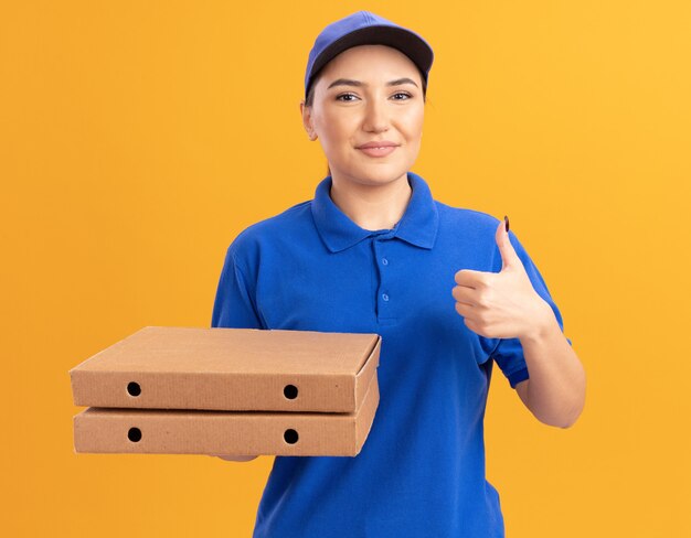 Feliz joven mujer de entrega en uniforme azul y gorra sosteniendo cajas de pizza mirando al frente sonriendo confiado mostrando los pulgares para arriba de pie sobre la pared naranja