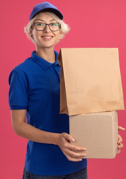 Feliz joven mujer de entrega en uniforme azul y gorra sosteniendo paquete de papel y caja de cartón sonriendo alegremente sobre pared rosa