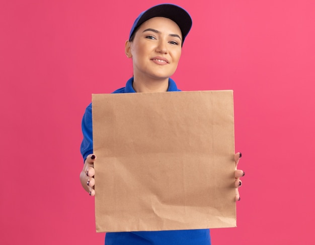 Feliz joven mujer de entrega en uniforme azul y gorra sosteniendo el paquete de papel mirando al frente sonriendo alegremente de pie sobre la pared rosa