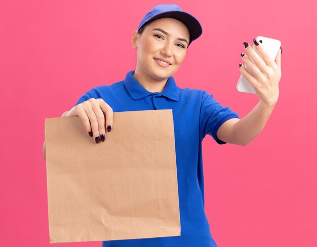 Feliz joven mujer de entrega en uniforme azul y gorra sosteniendo el paquete de papel que muestra el teléfono inteligente sonriendo alegremente de pie sobre la pared rosa